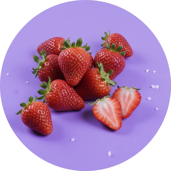 Ripe red strawberries with green tops, one halved to show interior, on purple surface with scattered white crystals.